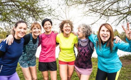 A line-up of six women standing outdoors dressed in exercise gear, smiling and with their arms around the person beside them.